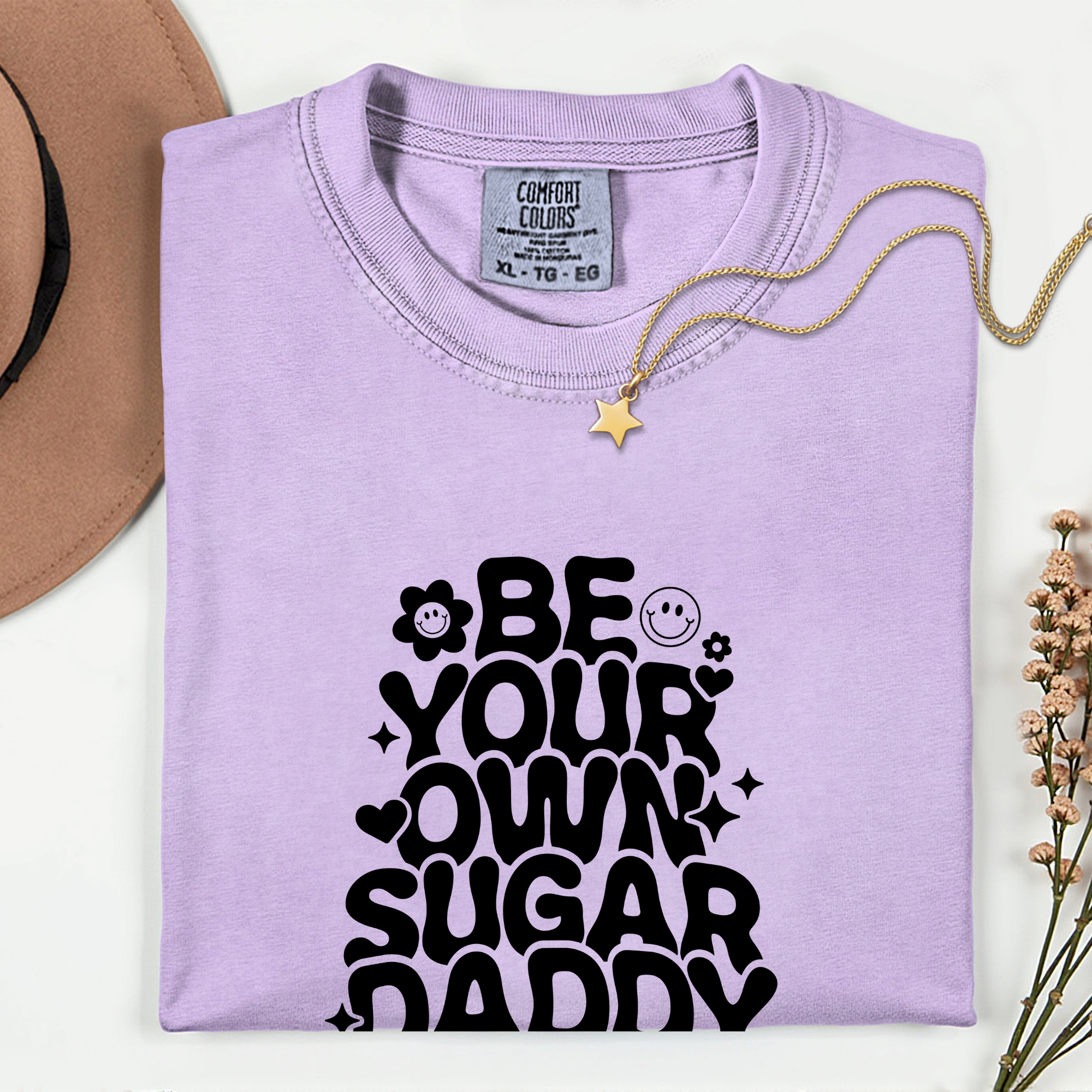 Folded purple sweatshirt with 'Be your own sugar daddy' text on a white background